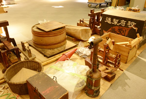 手前左の箱は、たばこ小売りの担い箱。隣の背の高い筒状のものは、羅宇屋の釜。奥の桶の右隣にある木製のものは、伊勢神宮の塩田の沼井の一部。
